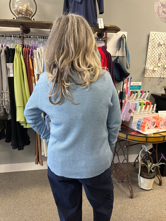 Person wearing a light blue sweater in a room with shelves and various items.