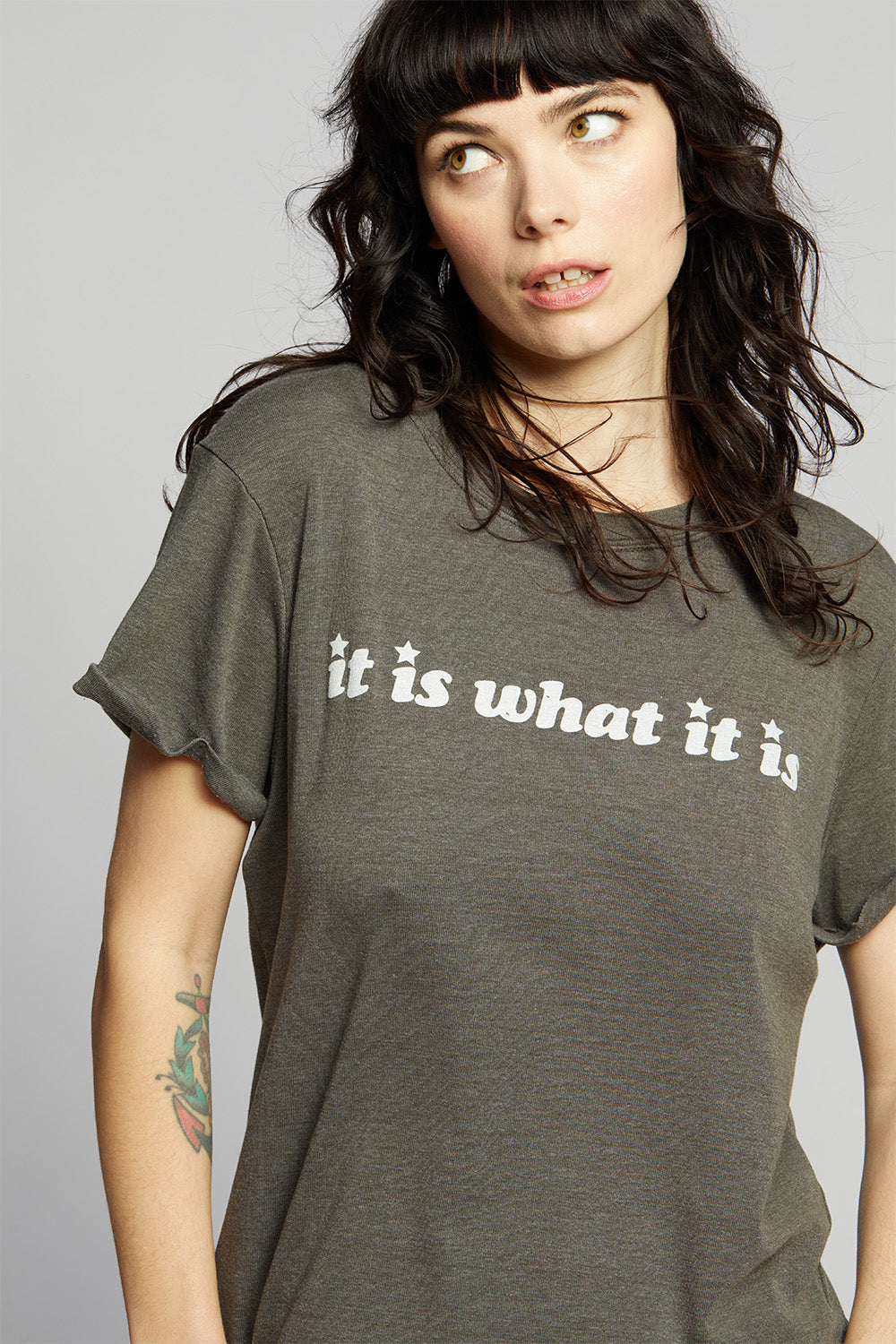 Woman wearing a gray t-shirt with 'it is what it is' text on a plain background