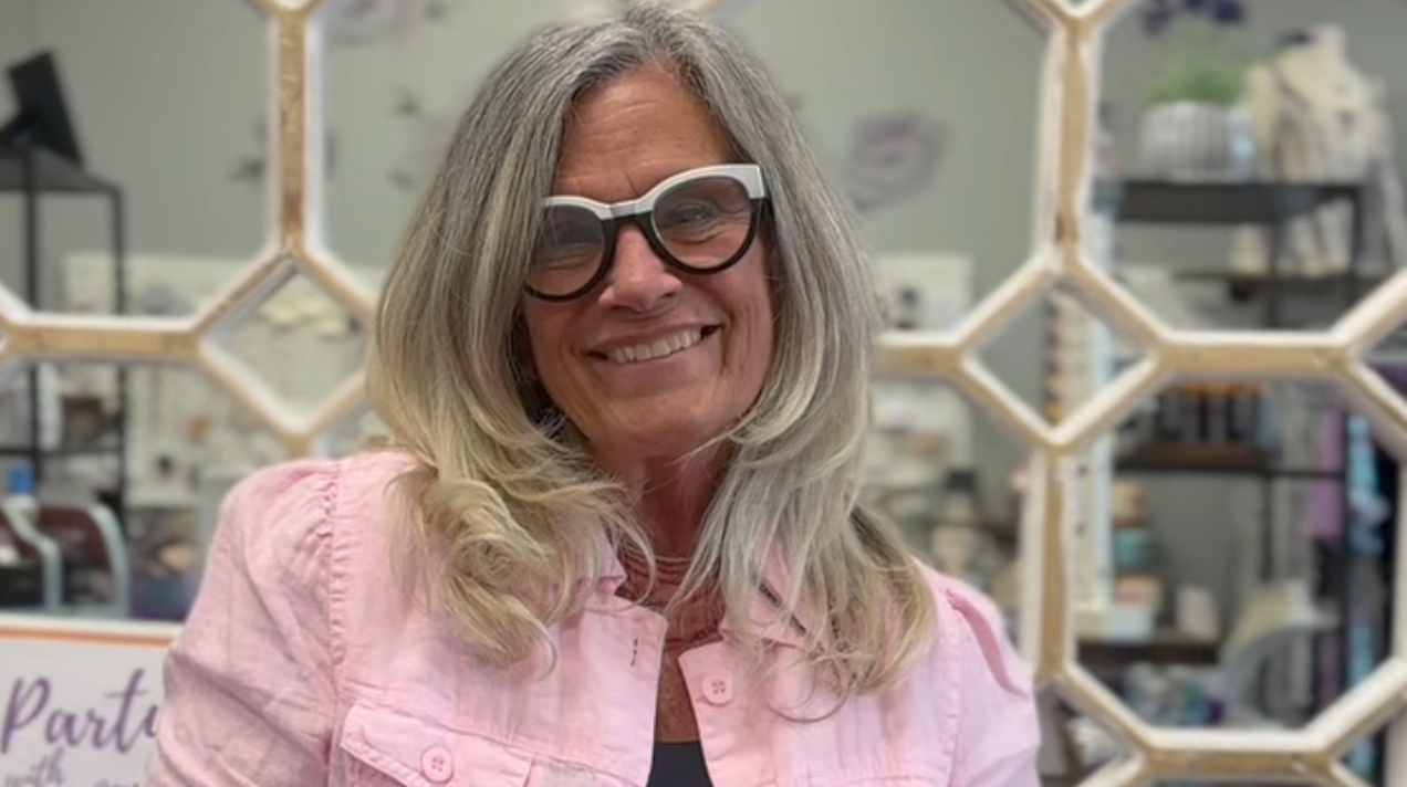 Woman with gray hair wearing white sunglasses and a pink shirt in a store setting.